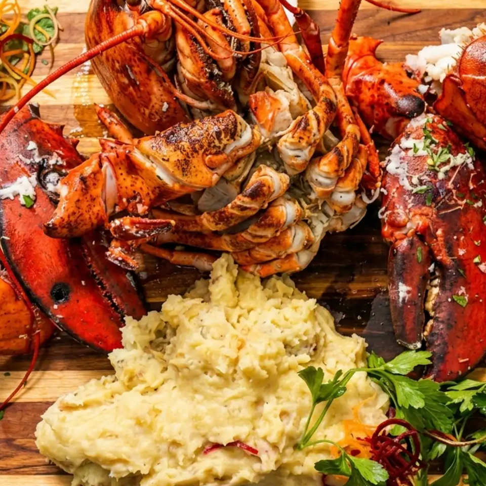 Overhead platter of lobster, crab legs, mashed potatoes, shredded vegetables, parsley, and bowls of melted butter on a wooden board.