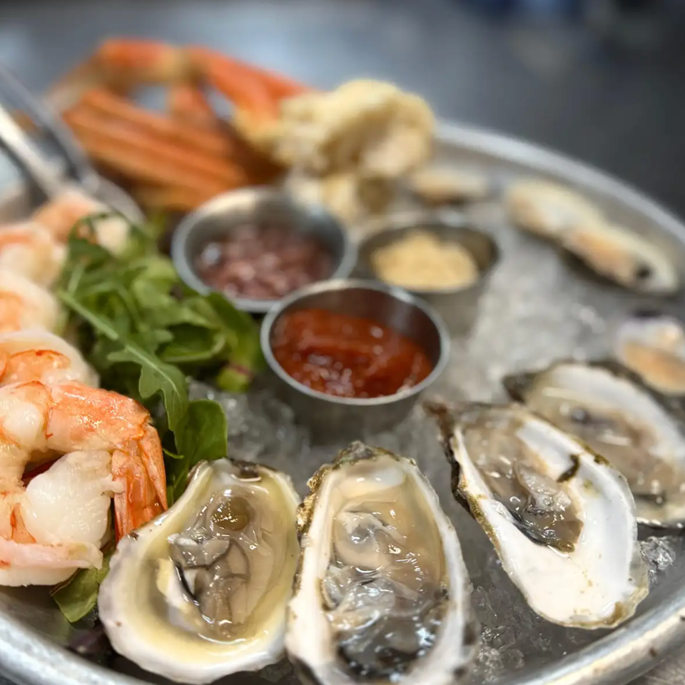 Seafood platter with shrimp, oysters, clams, mussels, greens, and dipping sauces served on ice.